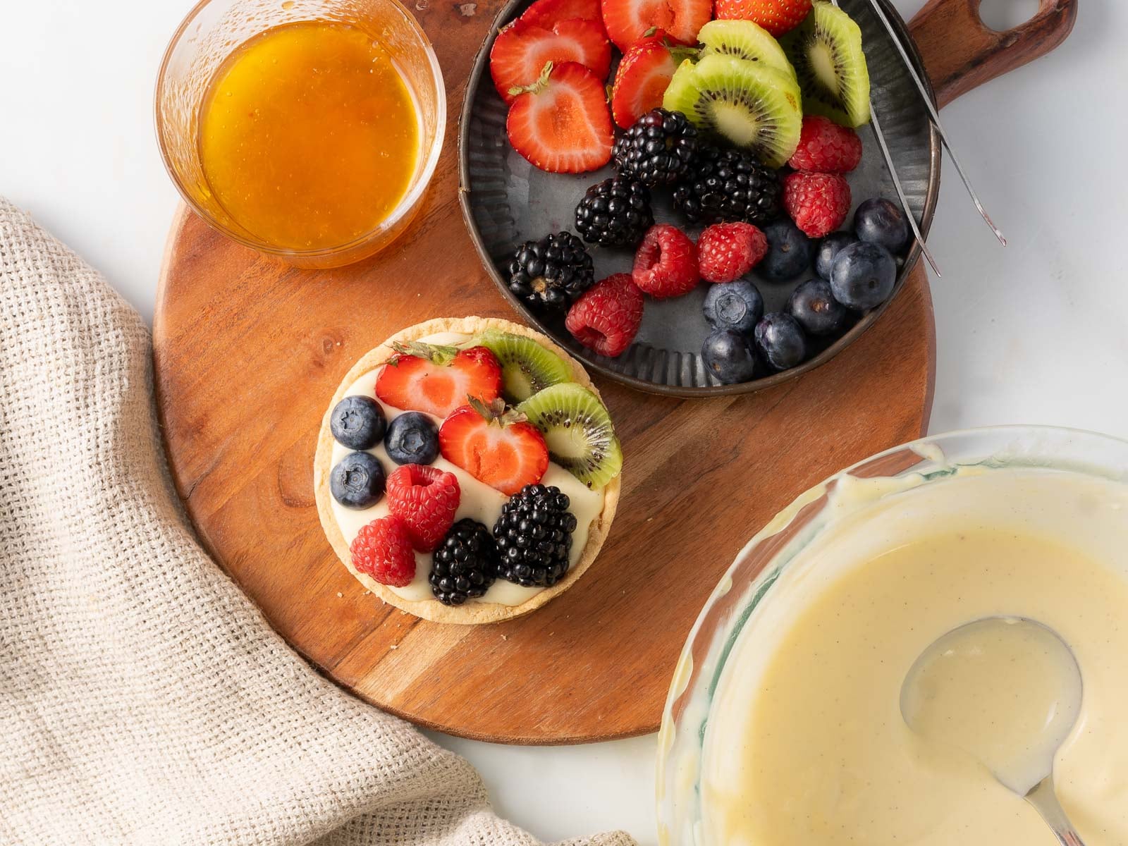 A tart topped with assorted berries and kiwi sits on a wooden board beside a bowl of fruit, custard, and a bowl of glaze.