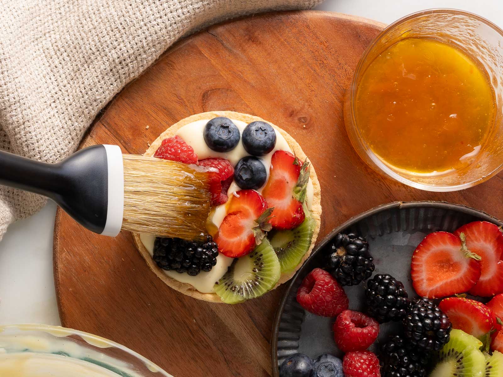 A pastry brush applies glaze to a fruit tart topped with strawberries, blueberries, blackberries, raspberries, and kiwi, on a wooden board beside a bowl of mixed berries and glaze.