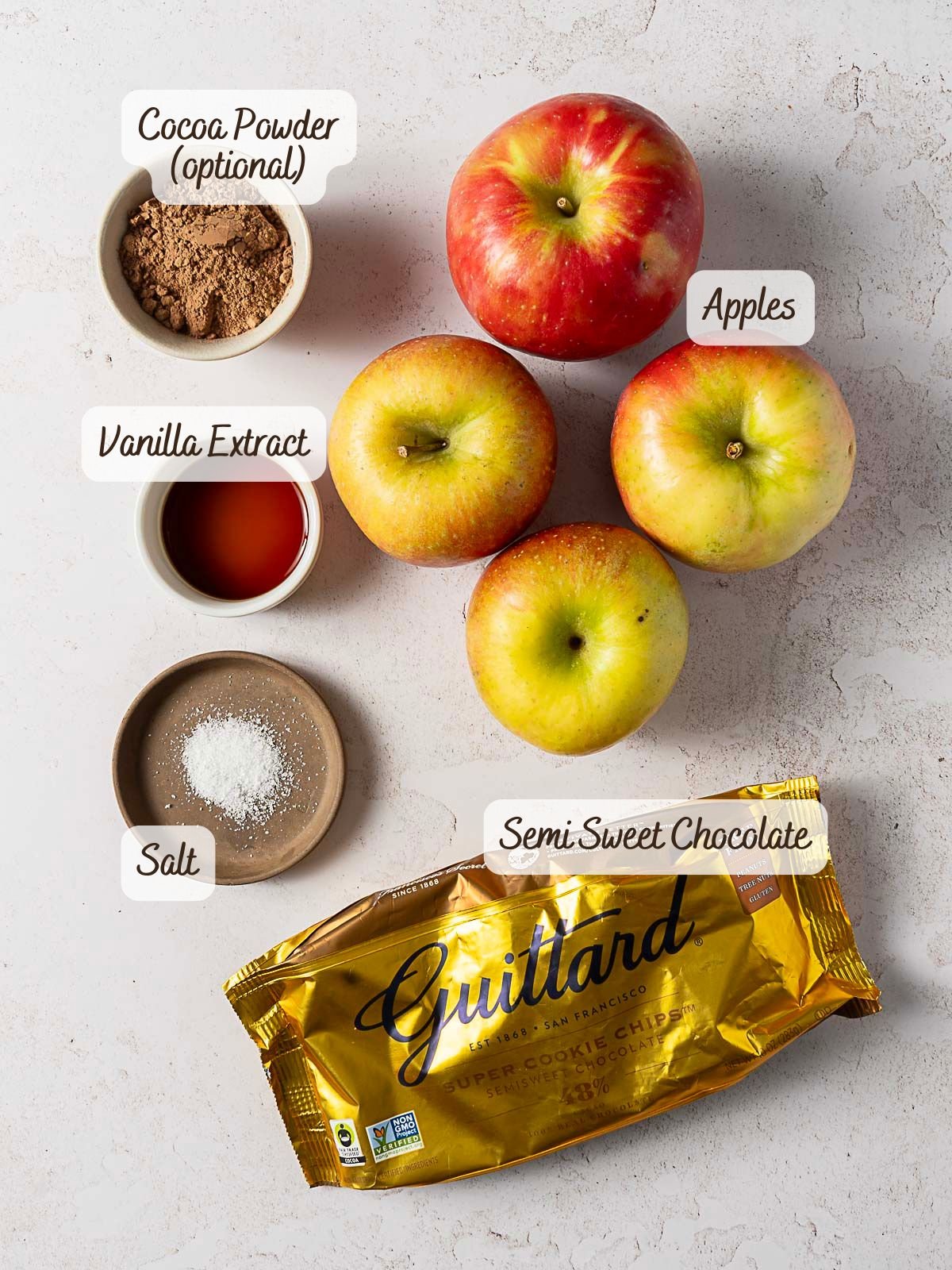 Four apples, a bowl of cocoa powder, vanilla extract, salt, and a package of Guittard semi-sweet chocolate chips-perfect for whipping up an easy no bake chocolate cake on a light surface.