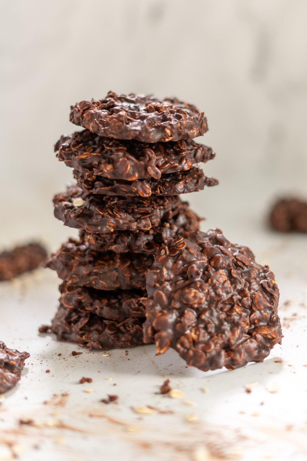 A stack of gluten-free chocolate oatmeal cookies is arranged on a light surface, with crumbs and a few scattered cookies in the background.