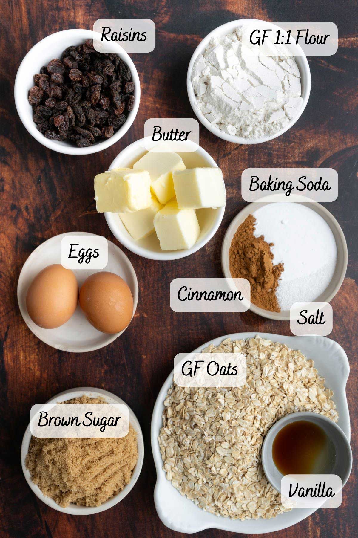 Top-down view of labeled ingredients for baking: raisins, gluten-free flour, butter, baking soda, cinnamon, eggs, salt, brown sugar, gluten-free oats, and vanilla on a dark surface.