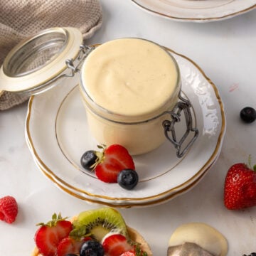 A glass jar of creamy pastry cream sits on a white plate, surrounded by fresh strawberries, blueberries, and sliced kiwi.