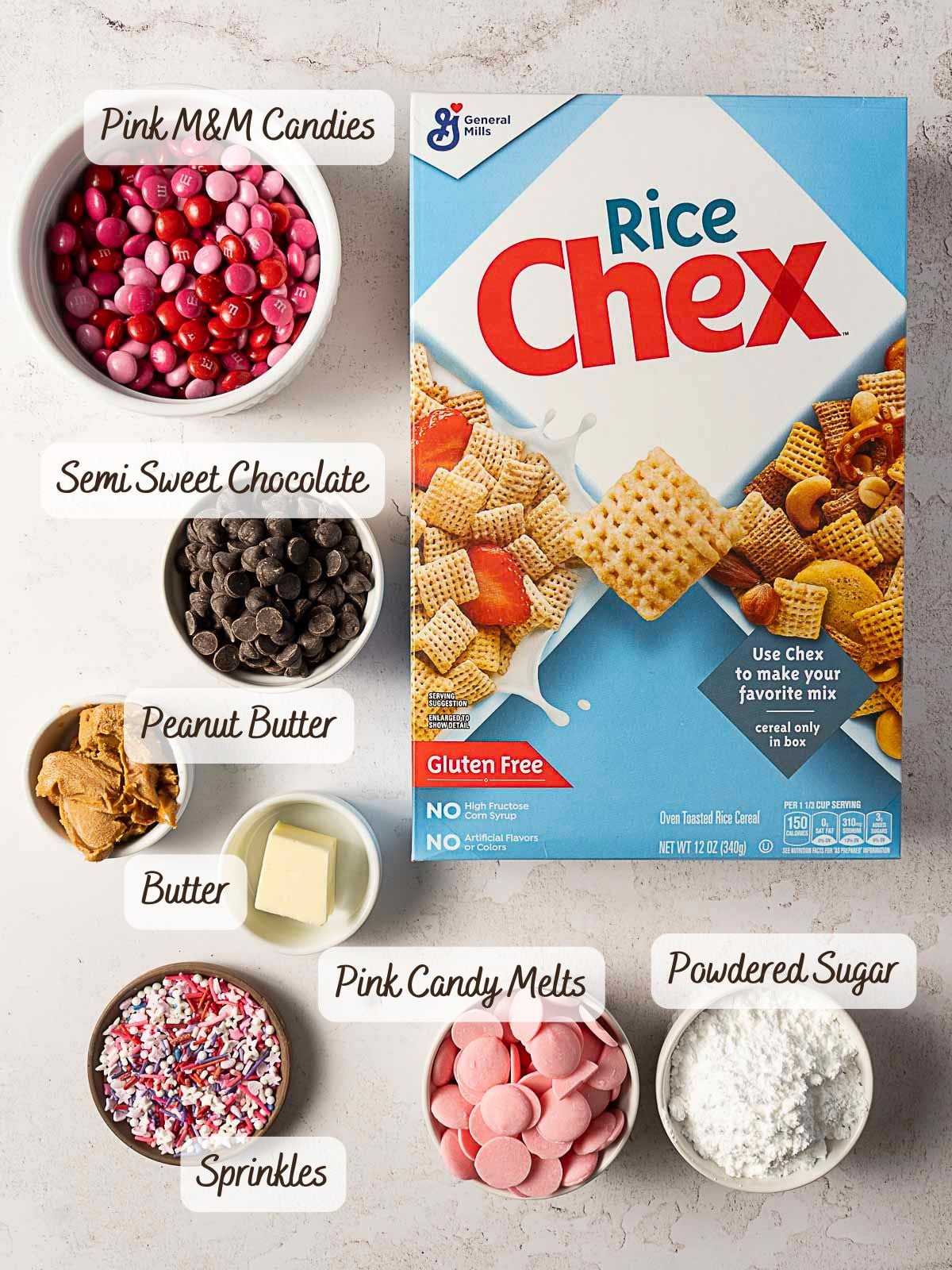 Ingredients for a dessert recipe are arranged, including Rice Chex cereal, pink M&M candies, semi-sweet chocolate, peanut butter, butter, pink candy melts, sprinkles, and powdered sugar.