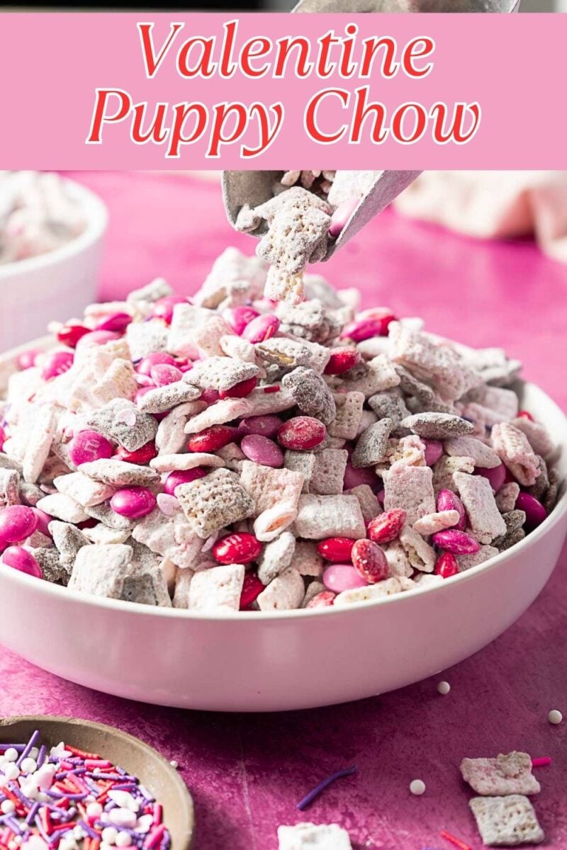A bowl of Pink Valentine Muddy Buddies snack mix with pink, red, and white candies being poured in, set on a pink tablecloth.