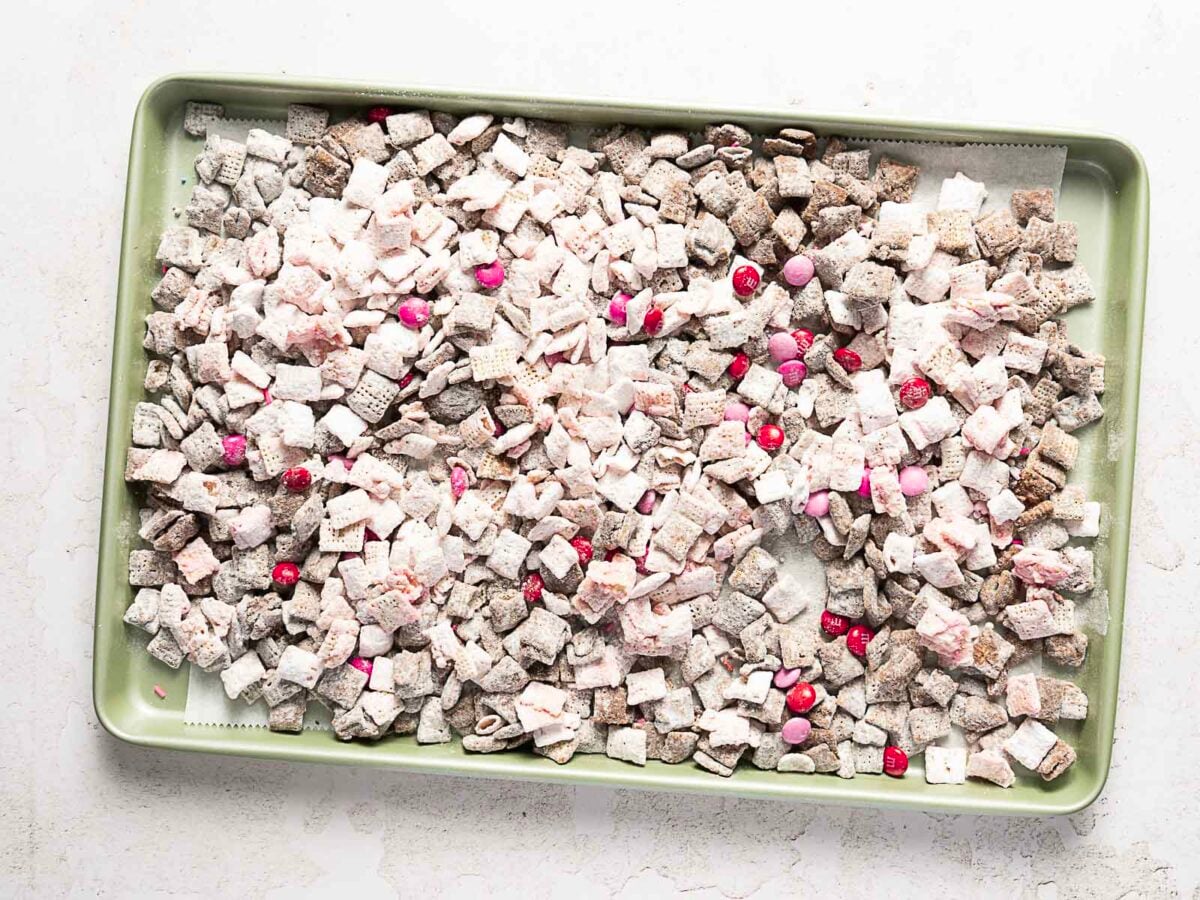A green baking tray filled with Chex cereal coated in powdered sugar, chocolate, and pink and red candies scattered throughout-perfect alongside a simple 3 ingredient cake for an easy, festive dessert spread.
