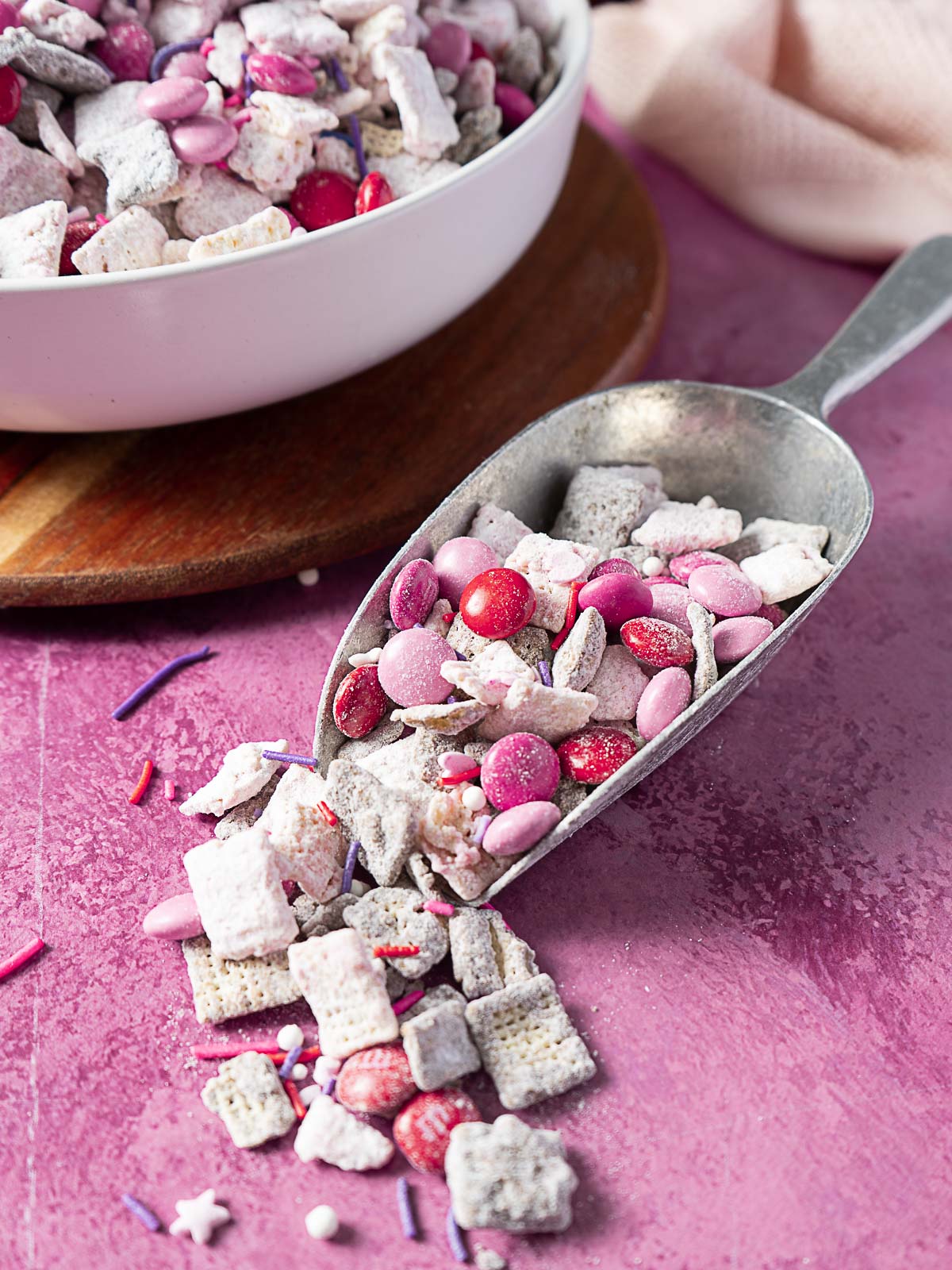 A metal scoop filled with pink, red, and white candy-coated snack mix rests on a pink surface next to a bowl of the same treat.