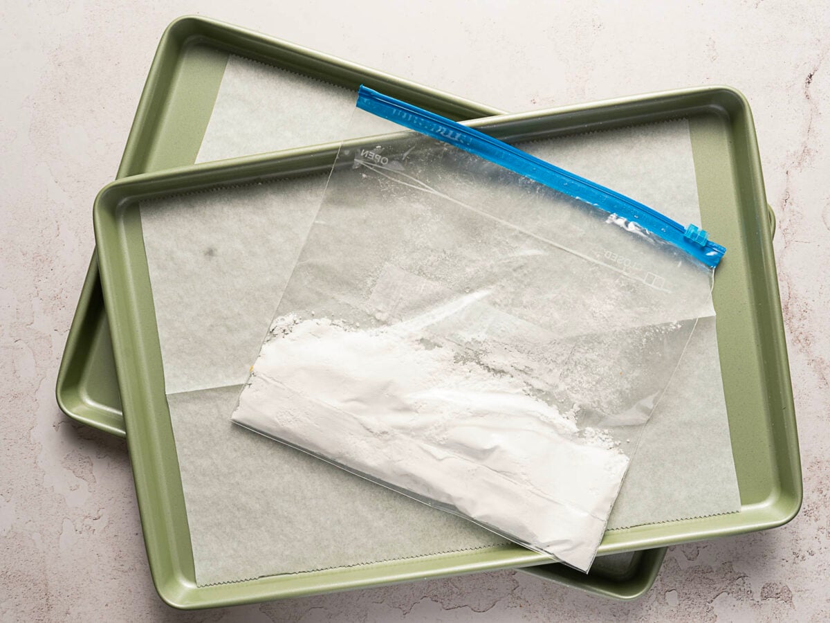 Two green baking sheets lined with parchment paper are stacked, with a clear plastic bag of white powder.