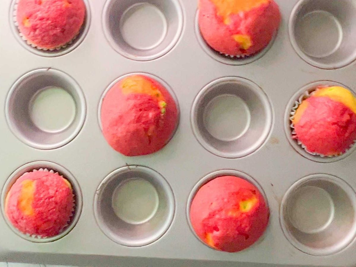 A muffin tin holds four pink and yellow muffins, made from a colorful cake mix, with five empty slots visible. The unevenly baked muffins sit in paper liners.
