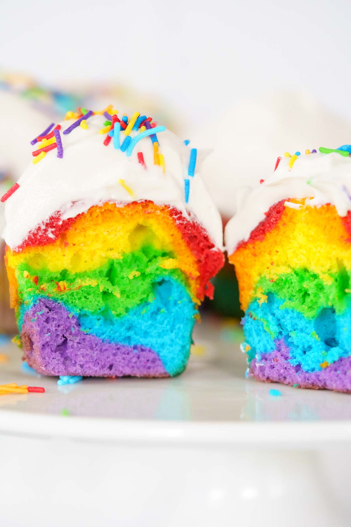 A sliced rainbow-colored cupcake with white frosting and multicolored sprinkles on a white plate&mdash;this vibrant treat is perfect for trying out your favorite Rainbow Cupcakes recipe.