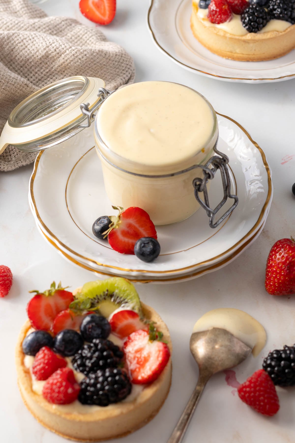 A jar of vanilla pastry cream on a plate beside a tart topped with fresh berries and kiwi; a spoon with custard rests nearby, surrounded by scattered fruit.
