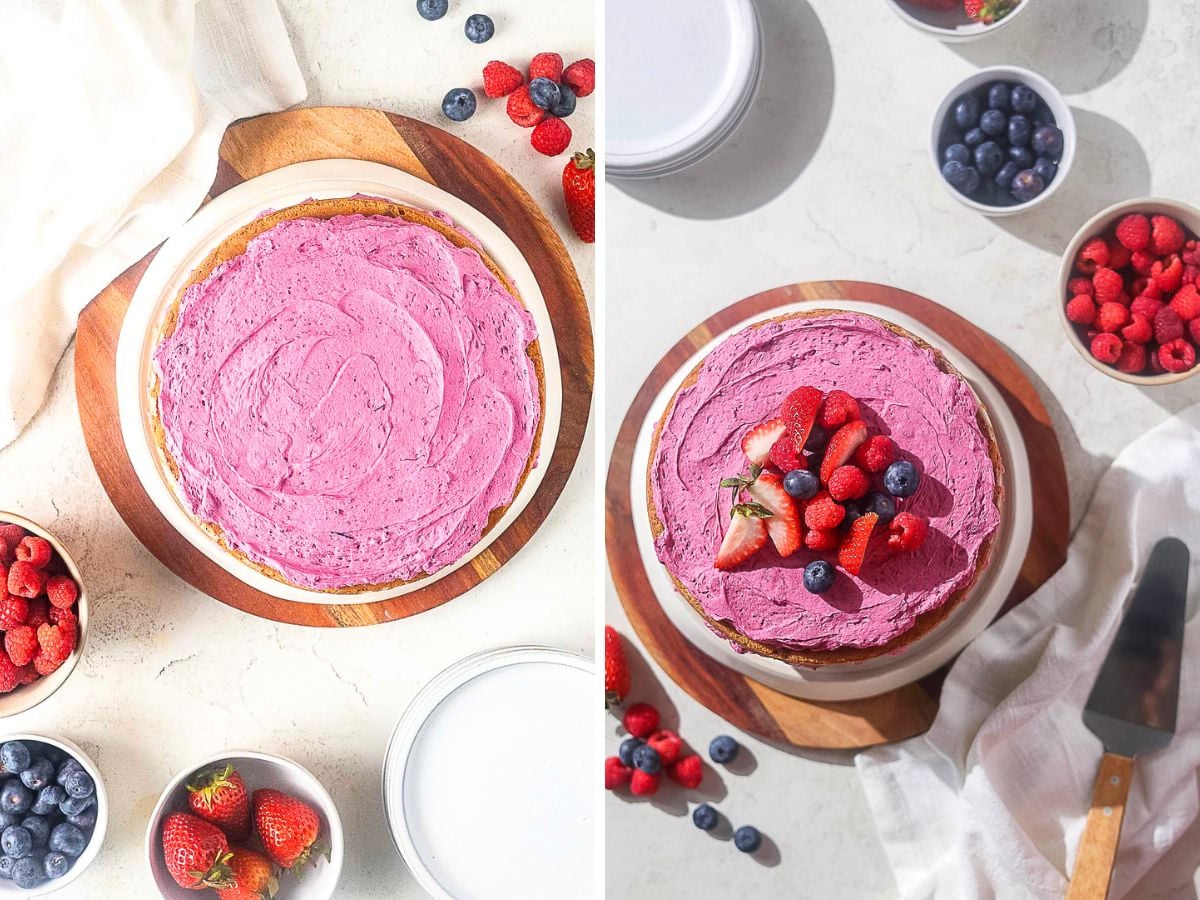 A gluten free blueberry cake with pink frosting sits on a wooden board, surrounded by bowls of strawberries and blueberries; some berries top the cake.