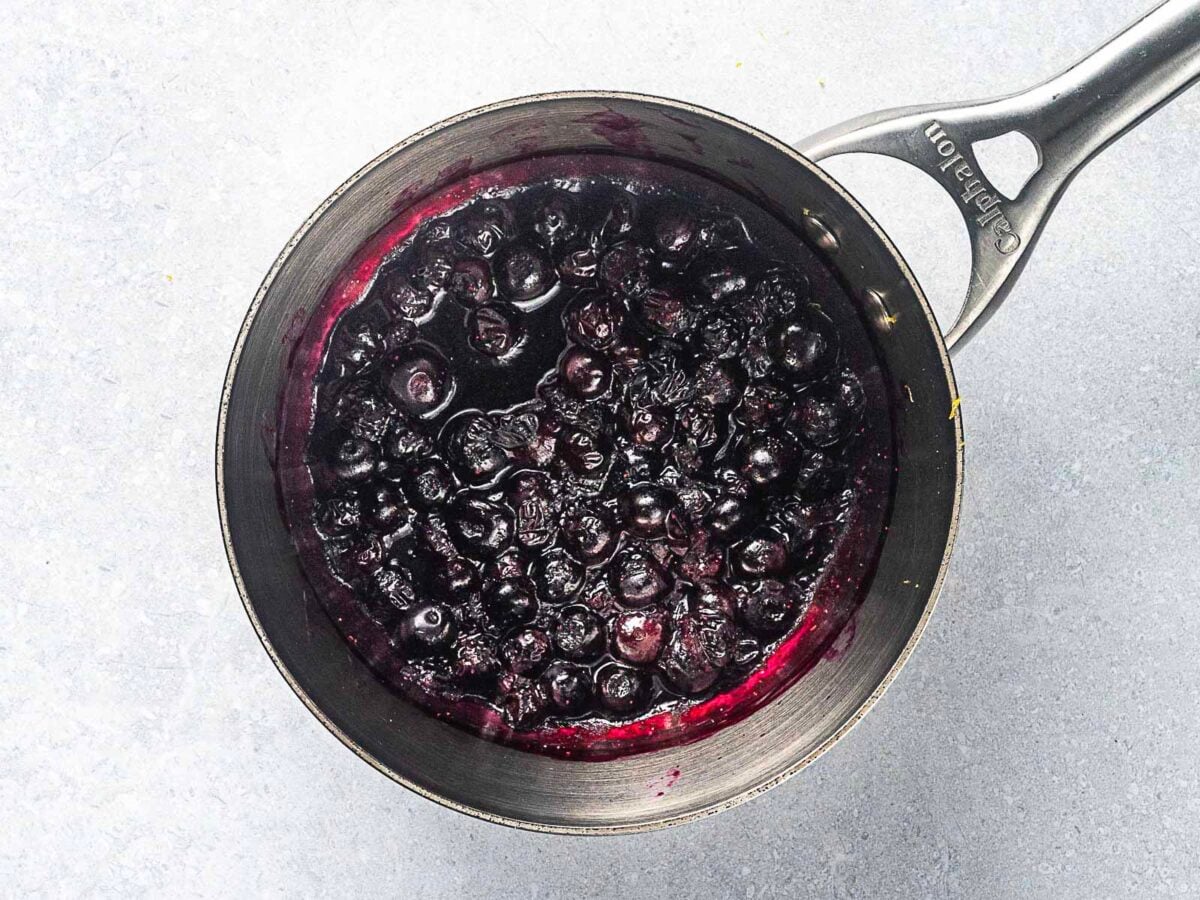 A stainless steel saucepan filled with cooked blueberries in sauce, perfect for making blueberry frosting, sits on a light gray surface.