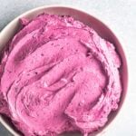 A bowl filled with smooth, bright pink blueberry frosting sits on a light gray surface.