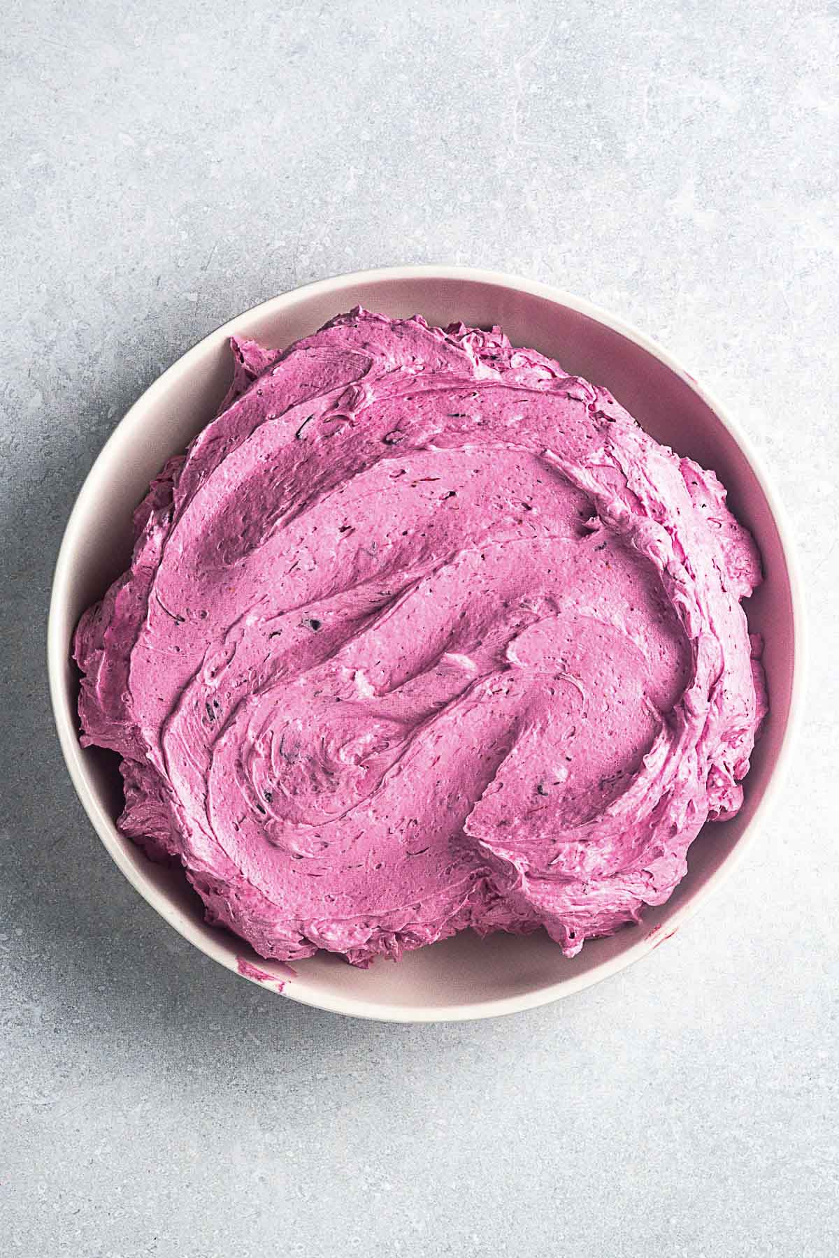 A white bowl filled with pink-purple blueberry frosting sits on a light gray surface.
