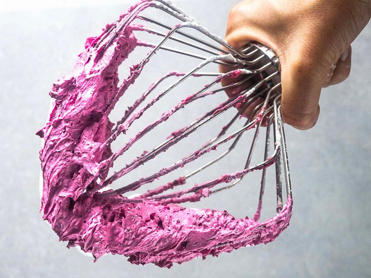 A hand holds a metal whisk covered in bright purple blueberry frosting against a plain grey background.