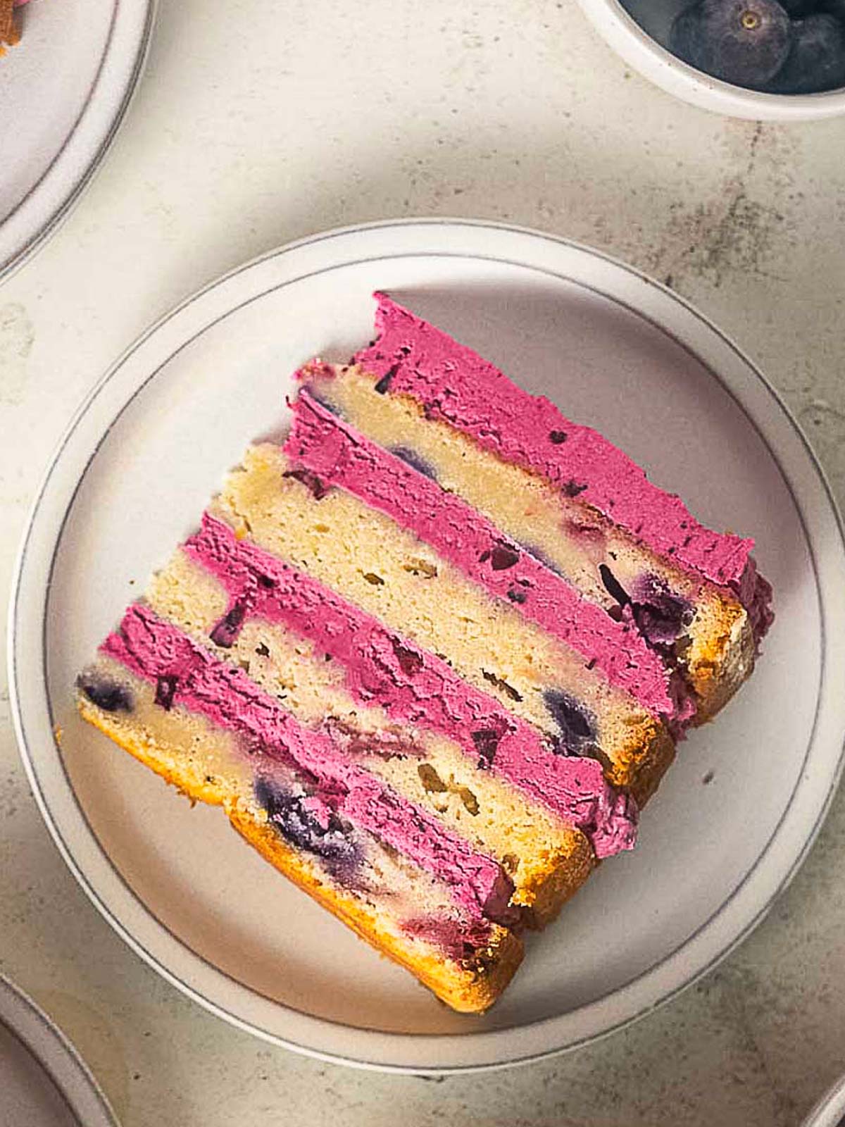 A slice of layered cake with purple blueberry frosting and visible blueberries on a white plate.