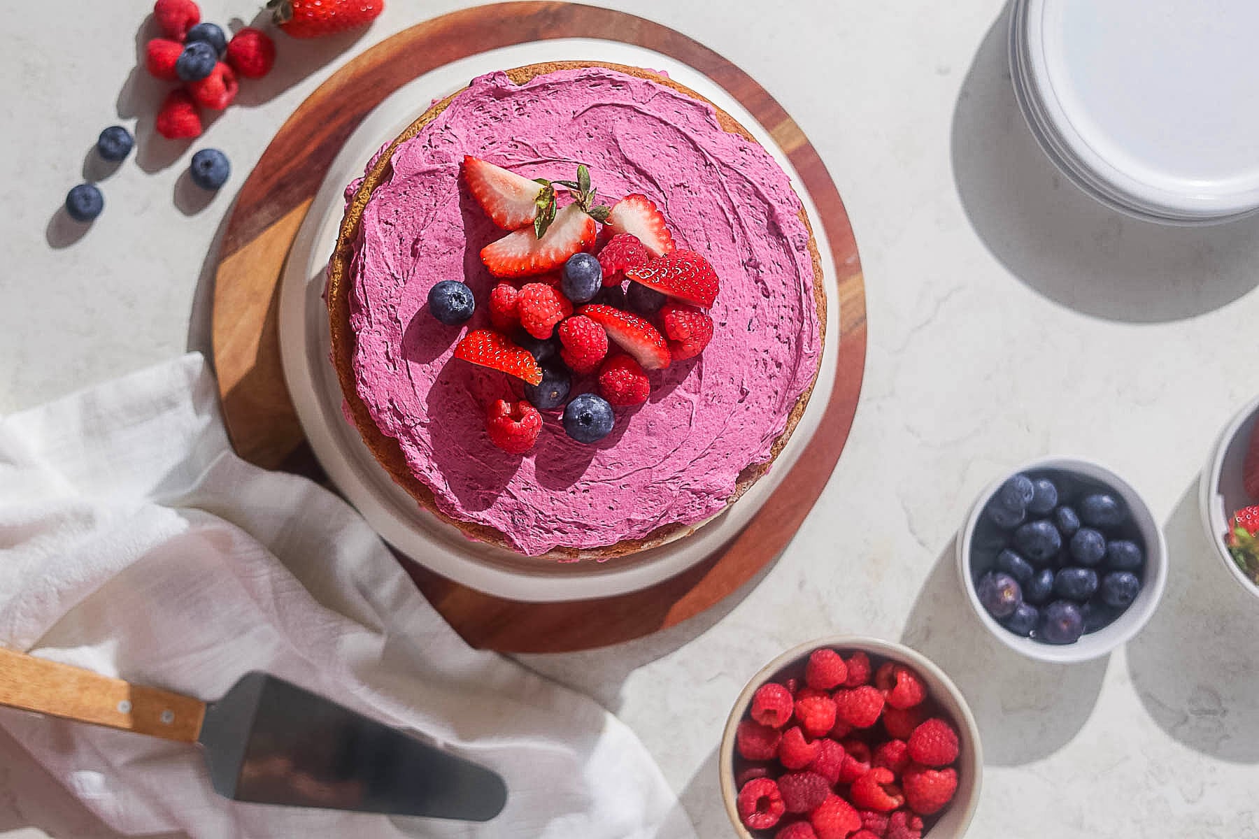 A Lemon Blueberry Cake on a cake stand with a bowl of berries.