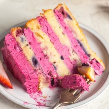 A slice of layered, gluten free blueberry cake with bright pink berry frosting and visible berry pieces, served on a white plate with a fork and a strawberry slice.