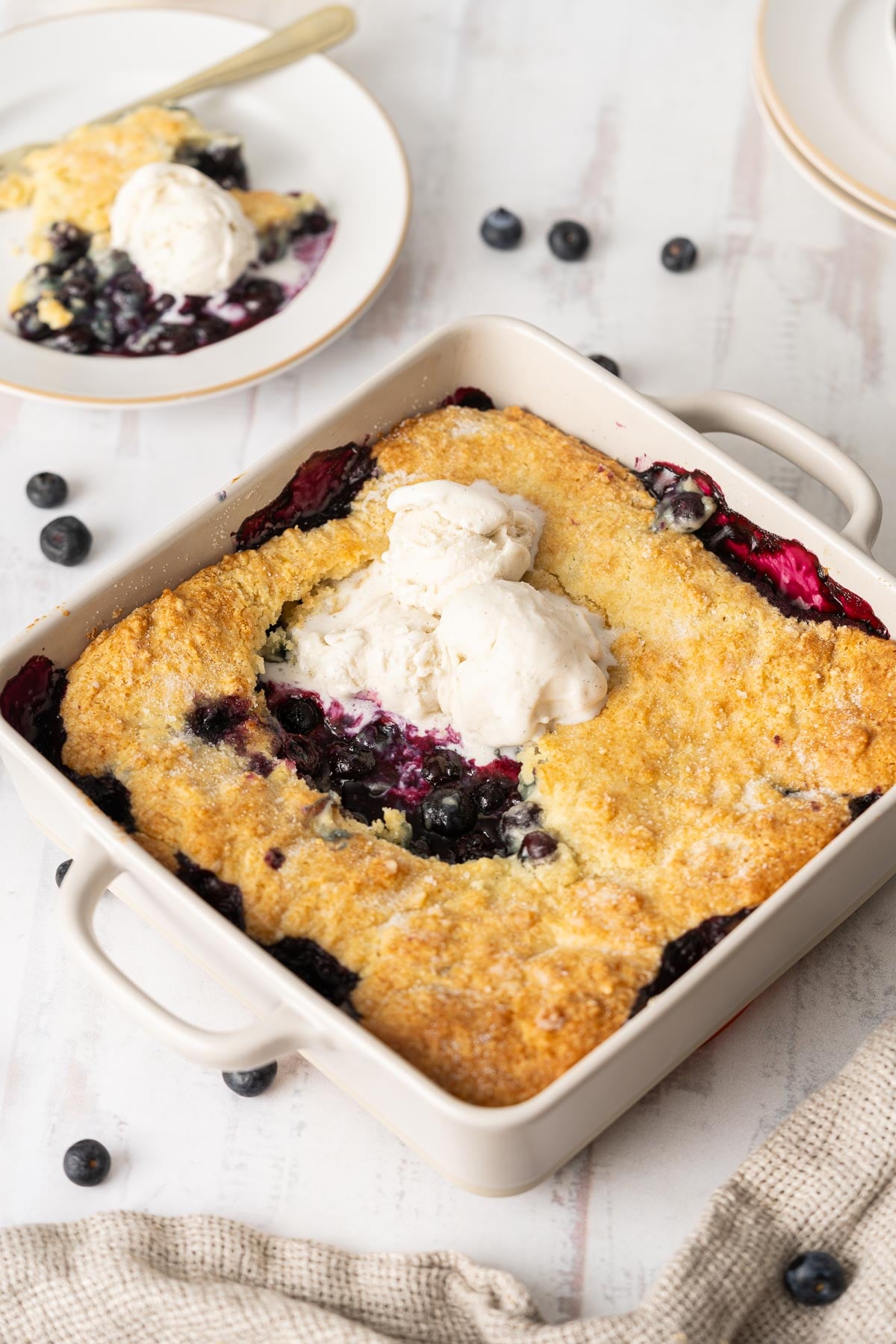 A square baking dish of gluten-free blueberry cobbler topped with vanilla ice cream, with a serving on a plate in the background. Fresh blueberries are scattered on the table.