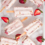 Rectangular strawberry popsicles with wooden sticks are arranged on a bed of ice, surrounded by fresh strawberries and strawberry halves.