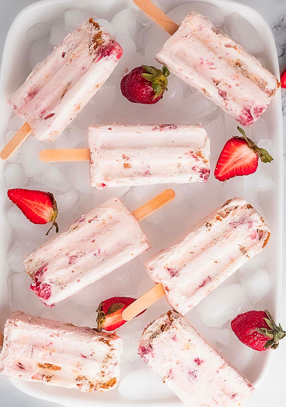 Several strawberry cheesecake popsicles are arranged on a tray of ice, surrounded by fresh strawberries and strawberry halves.