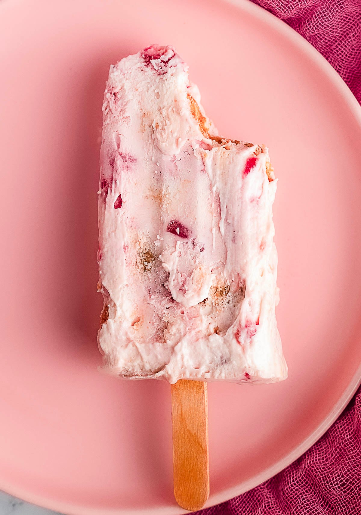 A creamy pink and white popsicle with visible fruit chunks and a bite taken out, resting on a pink plate.