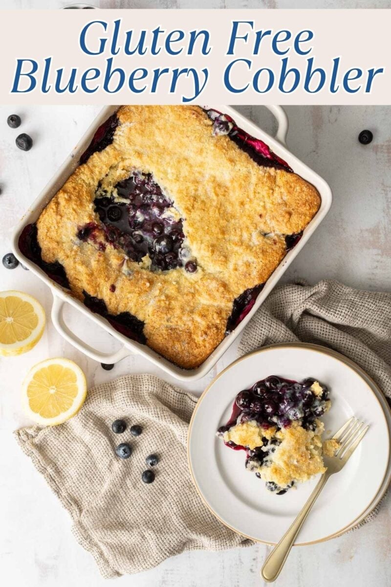 A baked gluten free blueberry cobbler in a square dish with a serving on a plate, surrounded by fresh blueberries and lemon slices on a white surface.