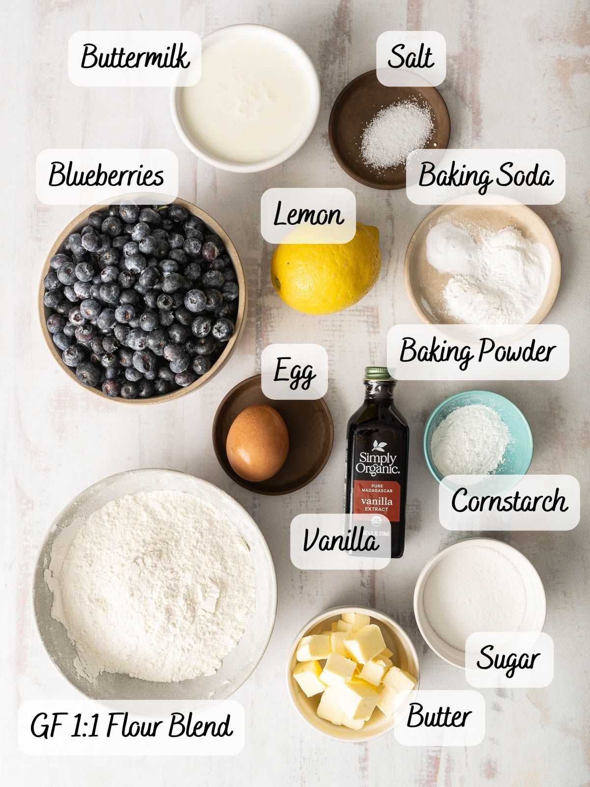 Overhead view of labeled ingredients for Gluten-Free Blueberry Cobbler: buttermilk, blueberries, lemon, egg, vanilla, gluten-free flour blend, butter, sugar, cornstarch, baking powder, baking soda, and salt.