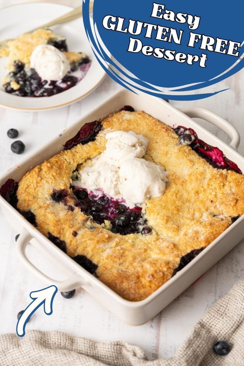 A baked gluten free blueberry cobbler topped with vanilla ice cream, served in a white baking dish with a portion on a plate in the background. Text reads, "Easy Gluten Free Dessert.