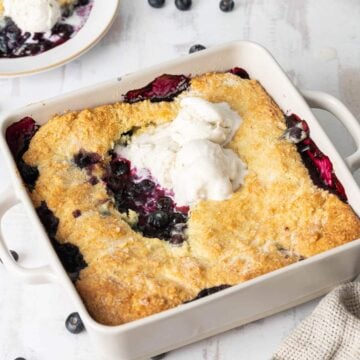 A baked blueberry cobbler in a square dish with two scoops of vanilla ice cream on top, sitting on a light-colored surface.