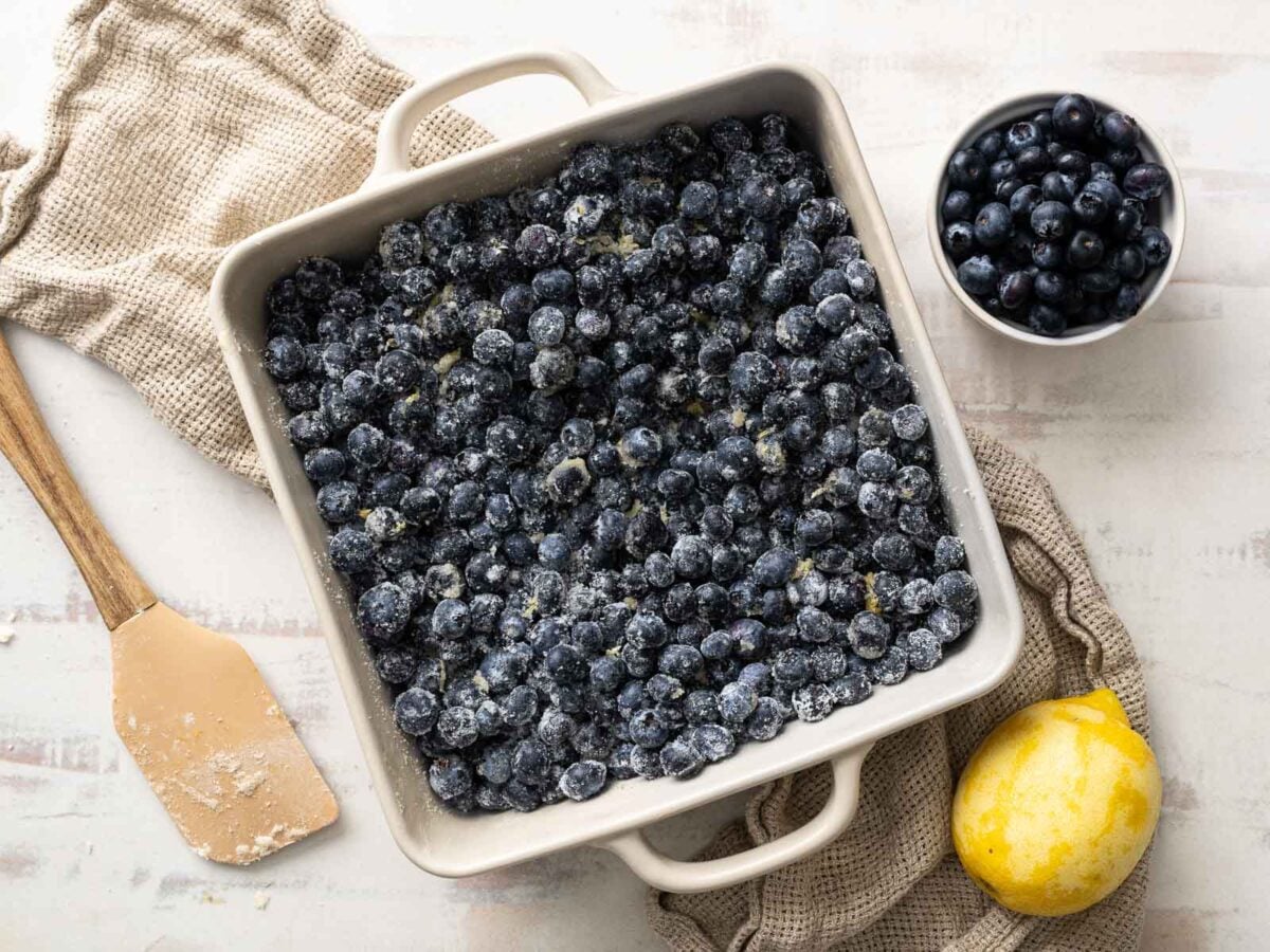 A square baking dish filled with sugared blueberries sits on a cloth, next to a bowl of blueberries, a lemon, and a spatula with flour-perfect for crafting a delicious gluten-free blueberry cobbler.