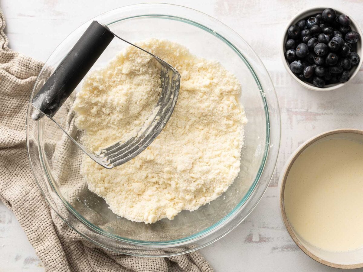 A glass bowl with gluten-free flour mixture and pastry blender, a bowl of blueberries, and a bowl of liquid batter on a light surface-perfect for making a delicious Gluten-Free Blueberry Cobbler.