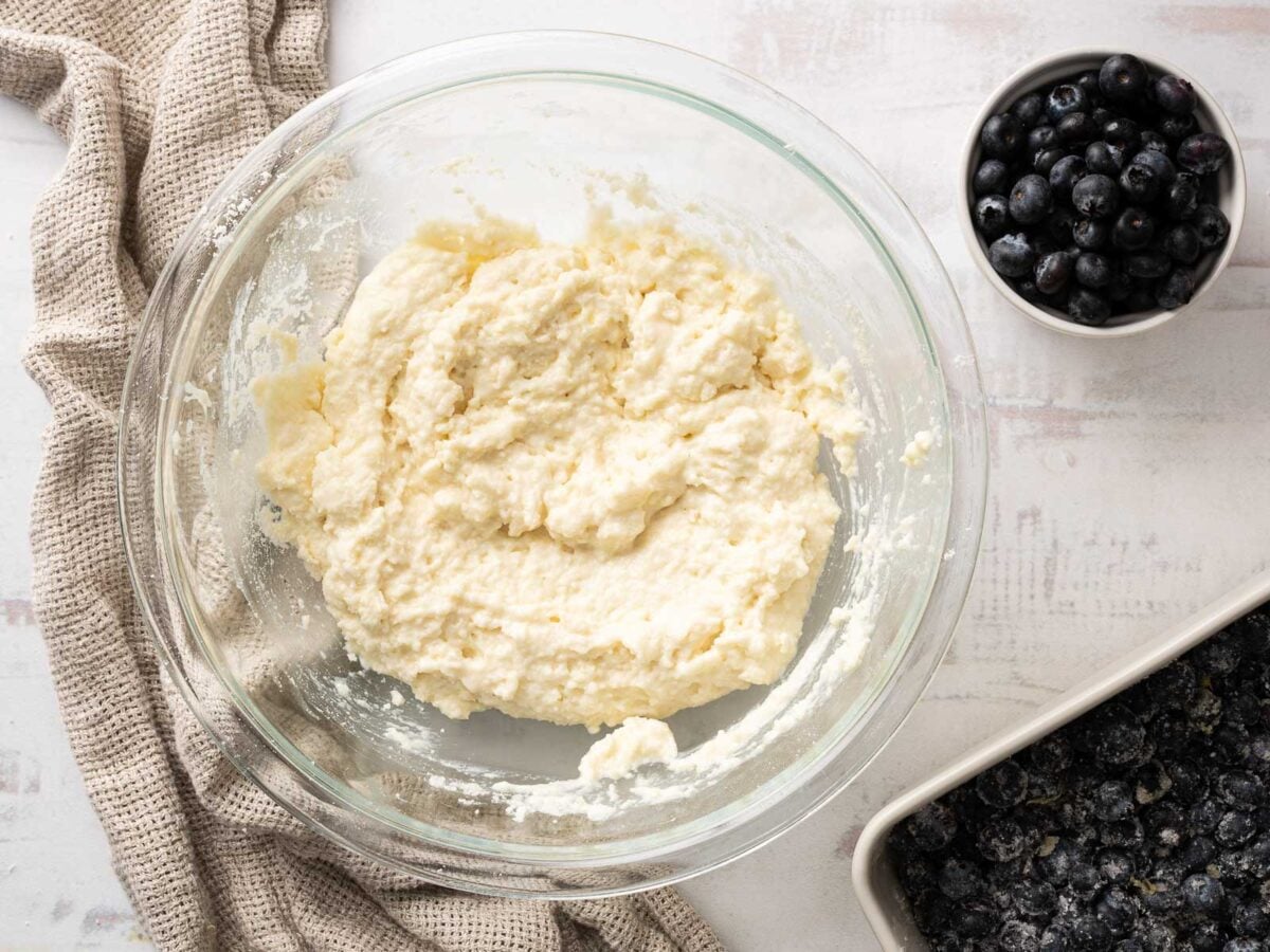 A glass bowl with mixed dough, a small bowl of fresh blueberries, and a baking dish with sugared blueberries on a light countertop-perfect for preparing a delicious Gluten-Free Blueberry Cobbler.