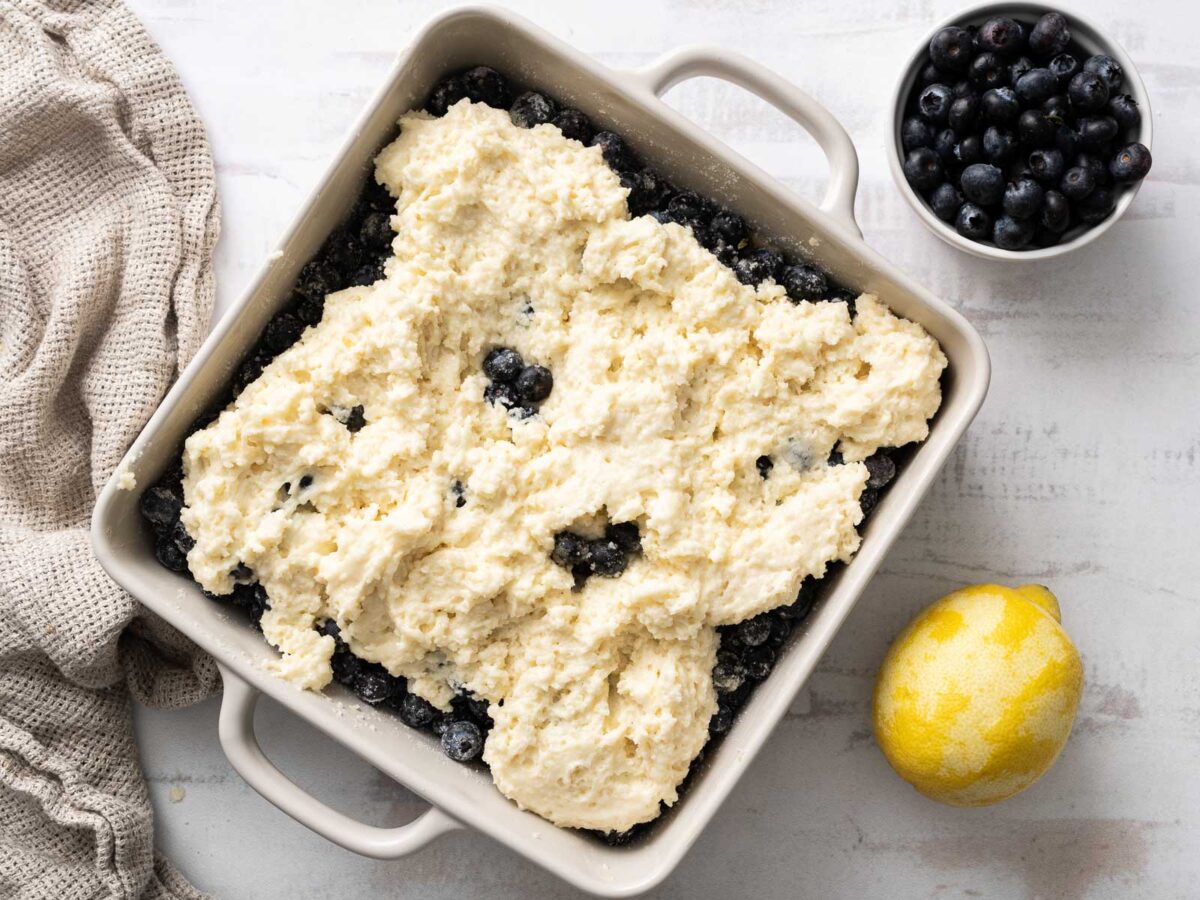Square baking dish with blueberries topped with dollops of biscuit dough, next to a cup of blueberries, a lemon, and a textured cloth on a light surface-a delicious gluten-free blueberry cobbler perfect for dessert.