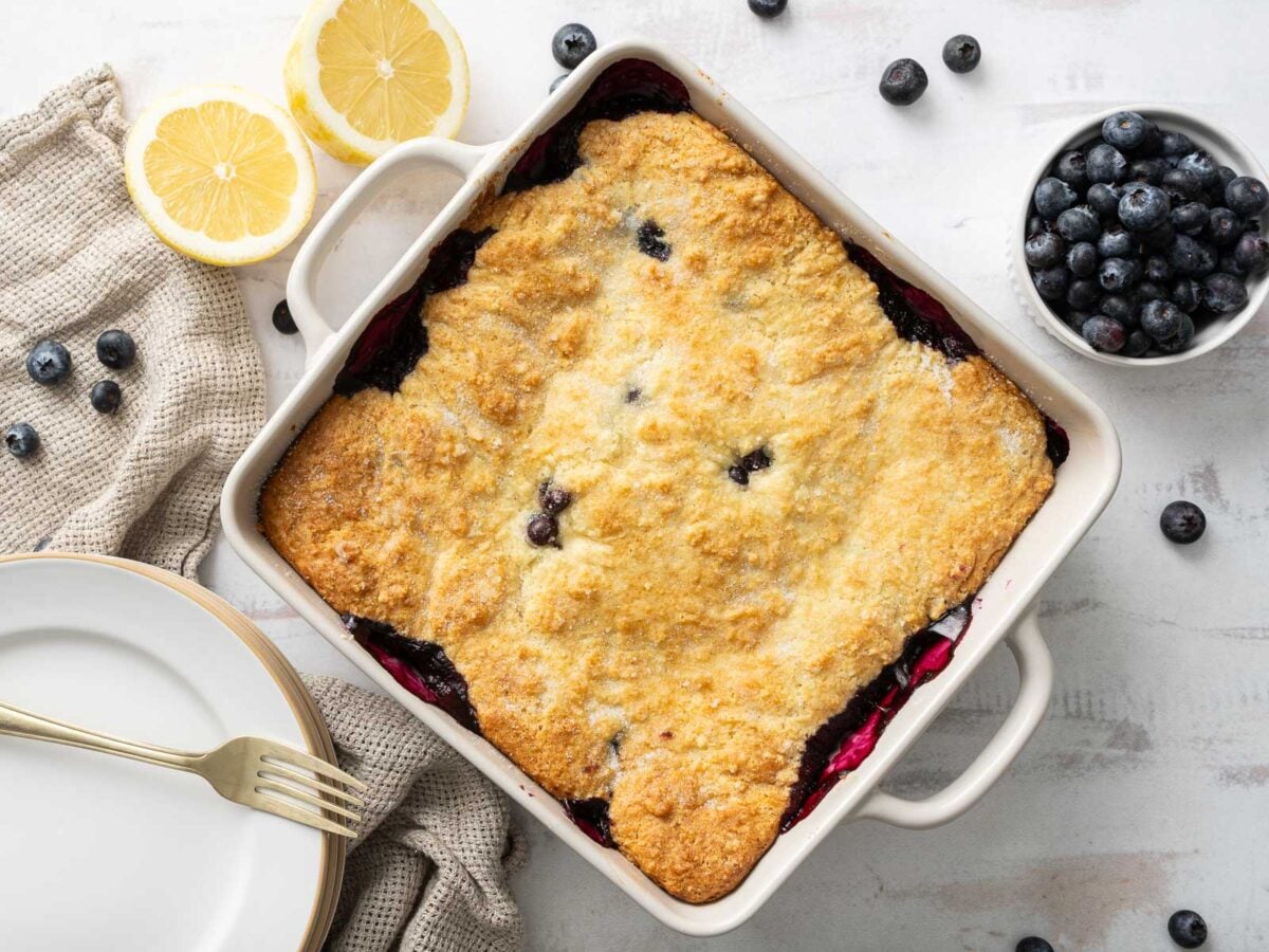 A baked gluten-free blueberry cobbler in a square dish, with a golden crust, surrounded by fresh blueberries, lemon halves, and plates with a fork.
