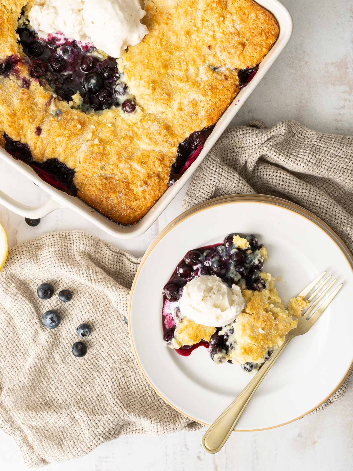 A serving of Gluten-Free Blueberry Cobbler with a scoop of vanilla ice cream on a plate, next to a baking dish filled with more cobbler and scattered blueberries.