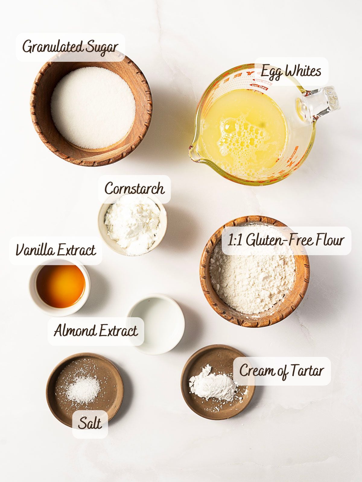 Top-down view of measured ingredients for Gluten Free Angel Food Cake in bowls and a glass jug, labeled as granulated sugar, egg whites, cornstarch, gluten-free flour, vanilla extract, almond extract, salt, and cream of tartar.