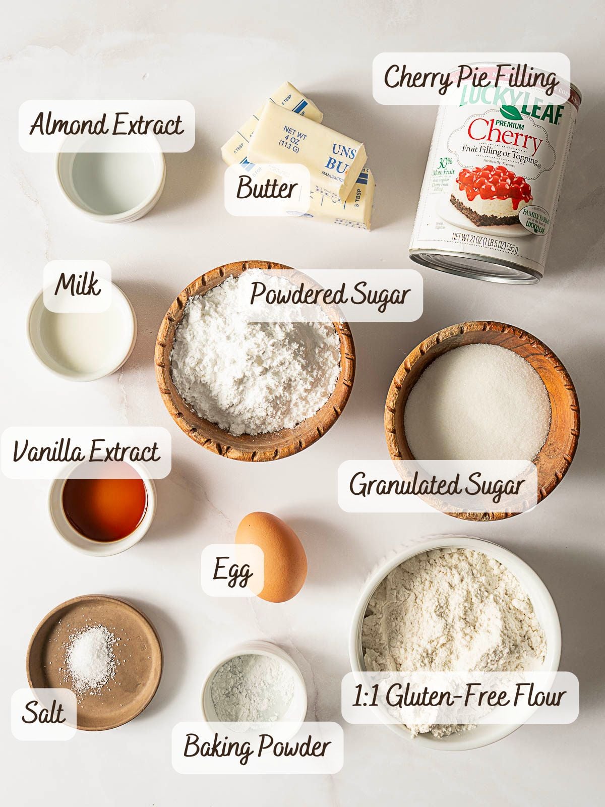 Various labeled baking ingredients for gluten free cherry pie bars, including butter, cherry pie filling, almond and vanilla extracts, milk, powdered and granulated sugar, an egg, salt, baking powder, and gluten-free flour.