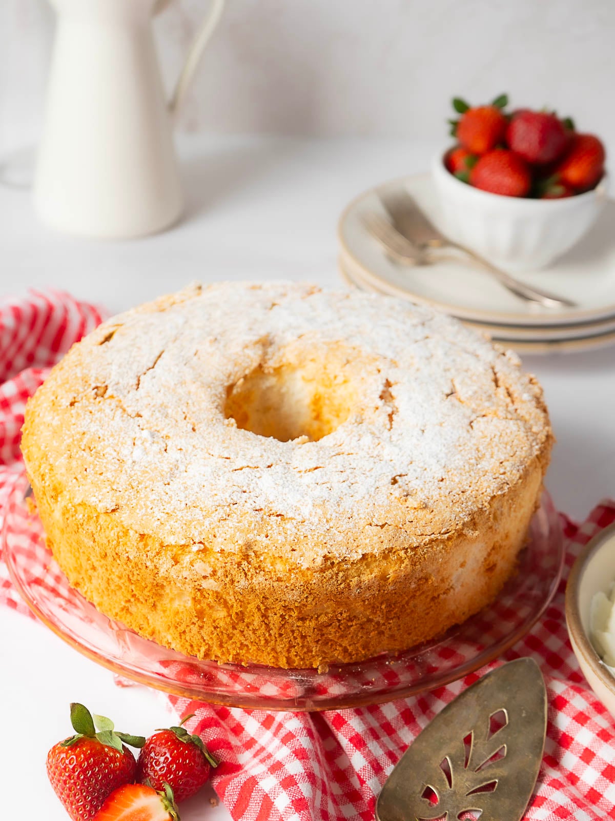 A golden-brown angel food cake dusted with powdered sugar sits on a glass plate next to fresh strawberries and a red checkered cloth.