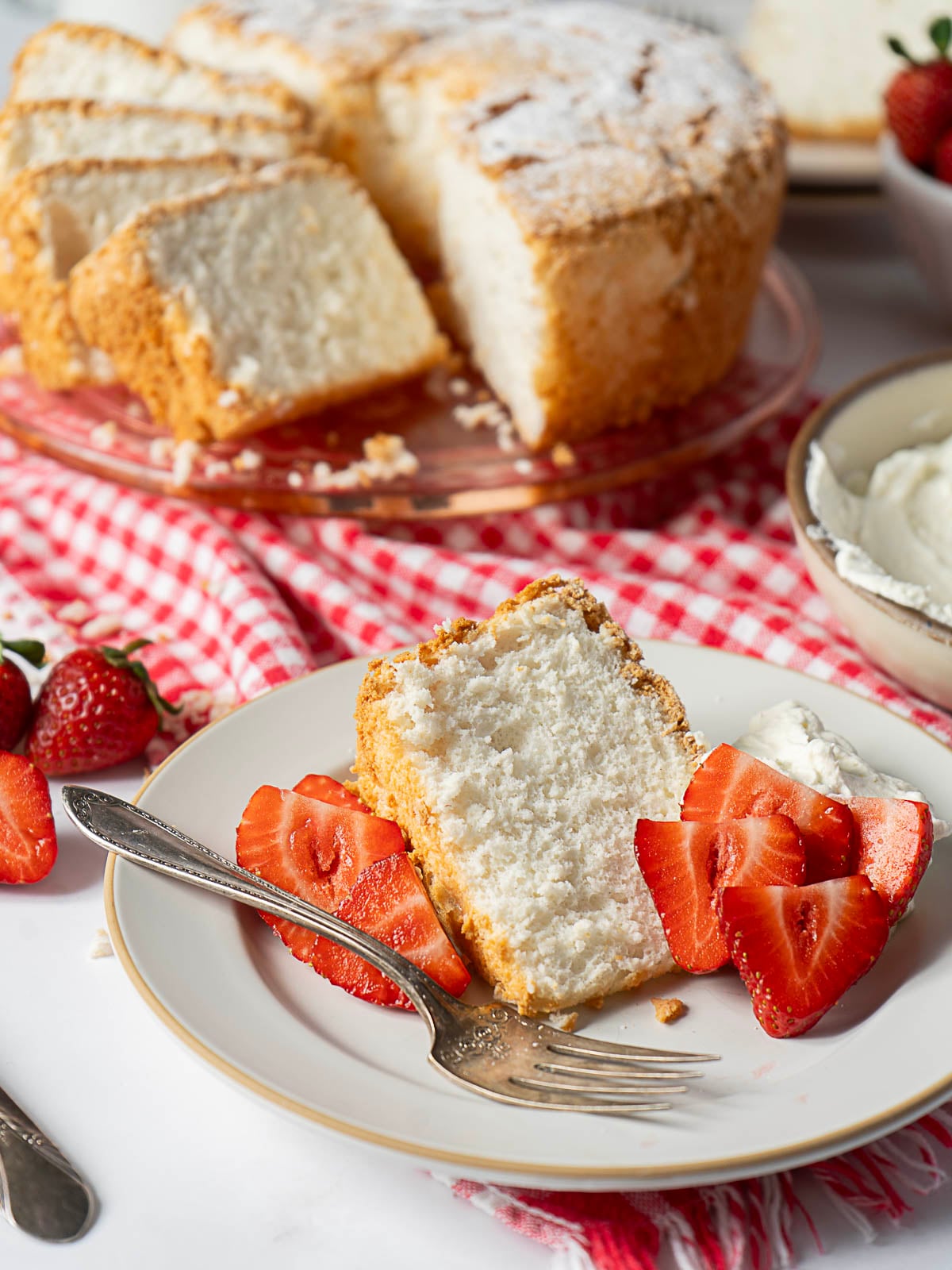 A slice of angel food cake with sliced strawberries and whipped cream on a plate, with the whole cake and more strawberries in the background.