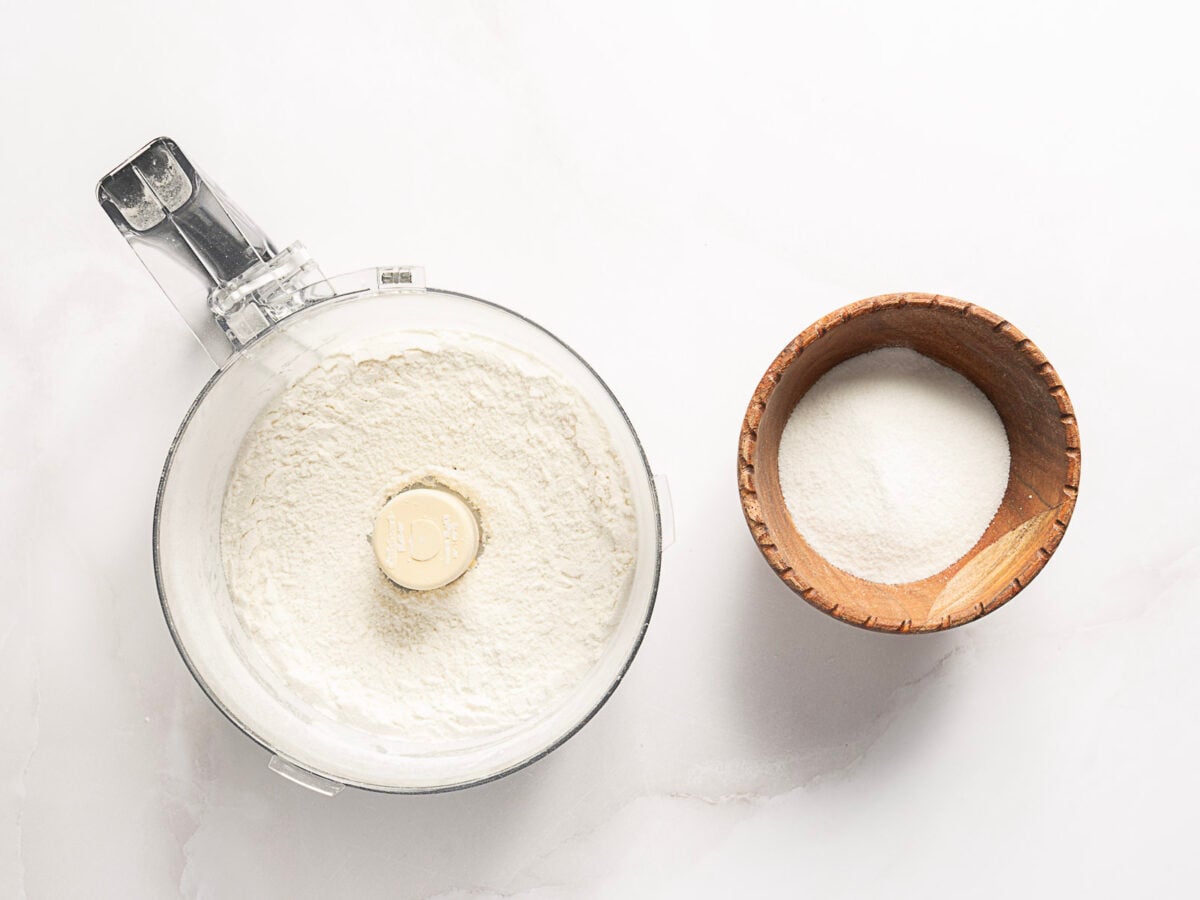 A food processor bowl filled with flour sits next to a small wooden bowl containing sugar on a white surface.