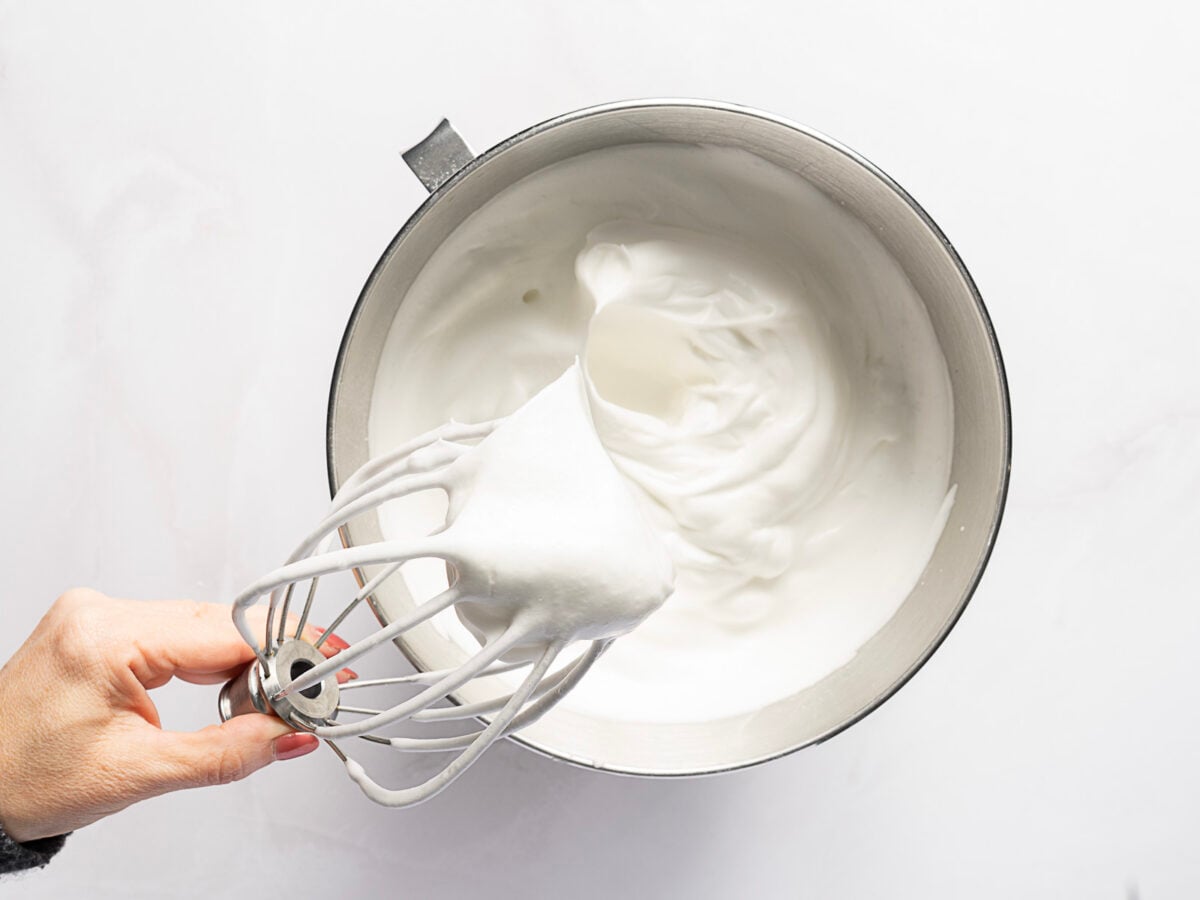 A hand holds a stand mixer whisk attachment with stiffly beaten egg whites above a mixing bowl containing more whipped egg whites.