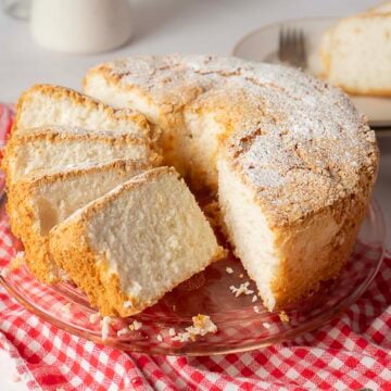 A round angel food cake with a light dusting of powdered sugar, partially sliced, sits on a glass plate atop a red and white checkered cloth.