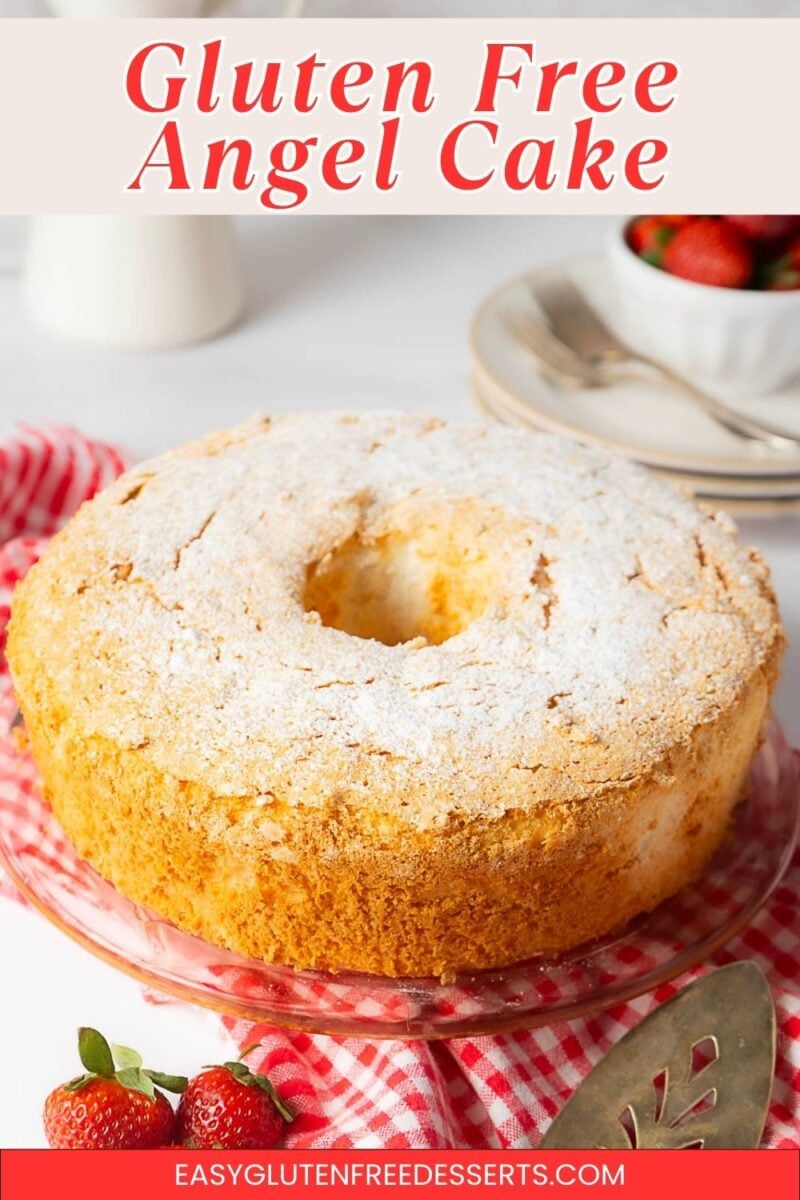 A Gluten Free Angel Food Cake dusted with powdered sugar sits on a glass cake stand, surrounded by strawberries and a red checkered cloth.