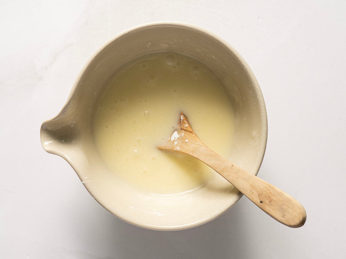 A ceramic mixing bowl filled with a pale yellow liquid batter and a wooden spoon resting inside it, placed on a white surface.