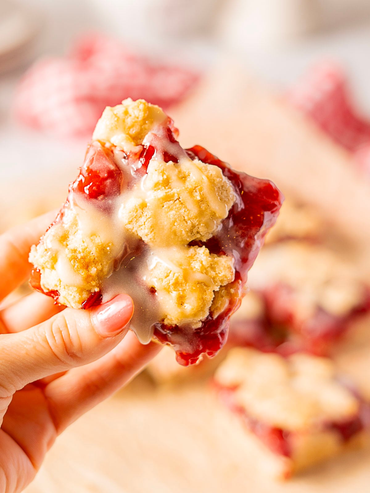 A hand holds a square cherry crumb bar topped with white icing; more bars are blurred in the background.