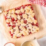 A square cherry crumble dessert topped with white icing sits on parchment paper beside a fork, a small jug of icing, and a red checkered cloth.