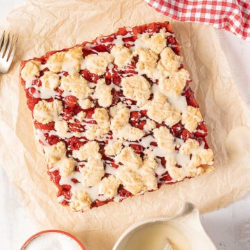 A square cherry crumble dessert topped with white icing sits on parchment paper beside a fork, a small jug of icing, and a red checkered cloth.