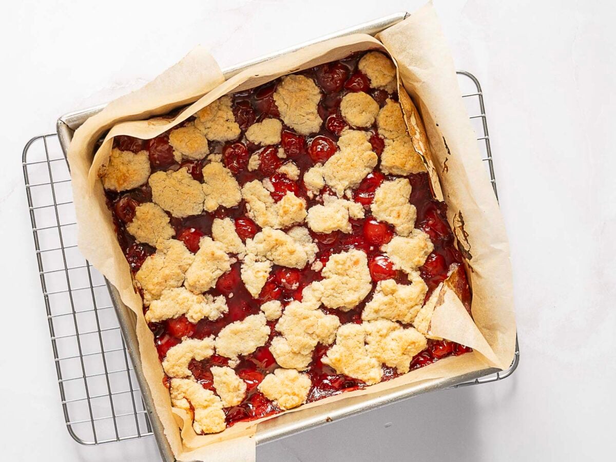 A square baking pan with parchment paper holds a cherry cobbler, featuring a golden, uneven biscuit topping over a layer of red cherry filling. The pan rests on a cooling rack.