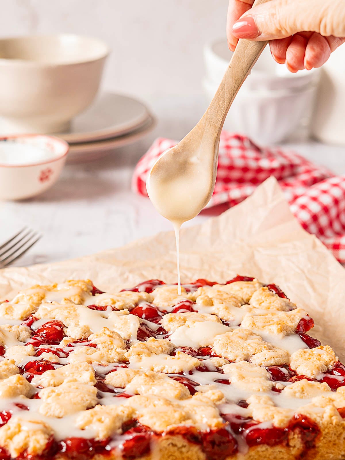 A hand drizzles white icing over a cherry crumb dessert on parchment paper, with dishes and a red-checked cloth in the background.